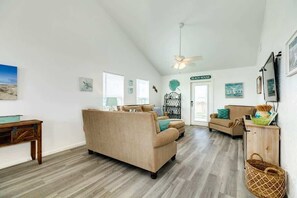 A spacious living room with a high ceiling, a ceiling fan, and a view of the beach through the door.
