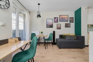 A modern living room with a dining area, a sofa, and a table with chairs. There are framed pictures on the wall and a window with curtains.