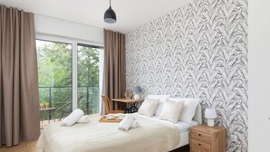 A hotel room with a large bed, a wooden nightstand, a table with a lamp, and a view of trees through the window.