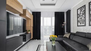 A modern living room with a large flat-screen TV, a black leather sofa, a coffee table, and a decorative vase with flowers.
