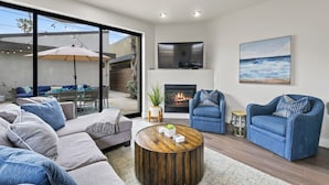 A modern living room with a fireplace, blue sofas, a coffee table, and a large window overlooking a patio area. - Pacific Beach Paradise by Cardo | Spacious Retreat (San Diego)