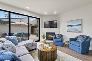 A modern living room with a fireplace, blue sofas, a coffee table, and a large window overlooking a patio area. - Pacific Beach Paradise by Cardo | Spacious Retreat (San Diego)