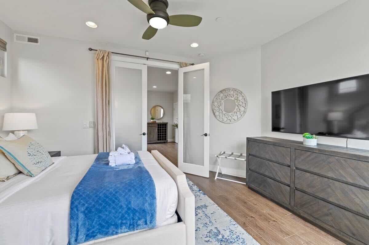 A modern bedroom with a bed, a TV, a ceiling fan, and a bathroom visible through an open door.