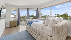 A bedroom with a large bed, a nightstand, a lamp, and a view of the outdoors. - Pacific Beach Paradise by Cardo | Spacious Retreat (San Diego)