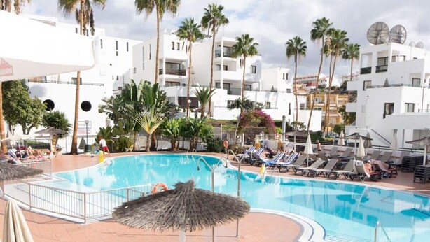 Una piscina con sillones de descanso y sombrillas, rodeada de palmeras y edificios blancos.