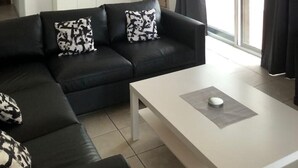 Una sala de estar moderna con un sofá de cuero negro, una mesa de centro blanca, un ventilador de techo, un espejo grande y una puerta corrediza de vidrio con cortinas.