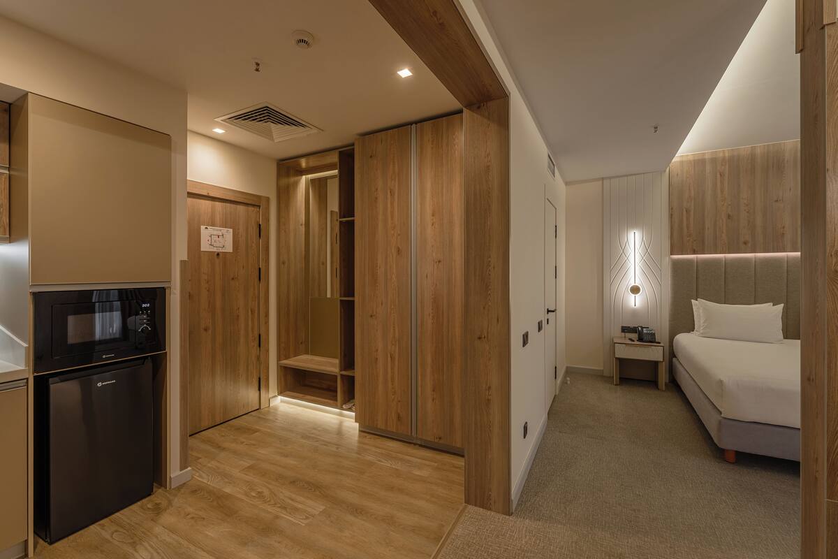 A modern hotel room with a wooden wardrobe, a built-in microwave, a small bedside table with a lamp, and a bed with white bedding.