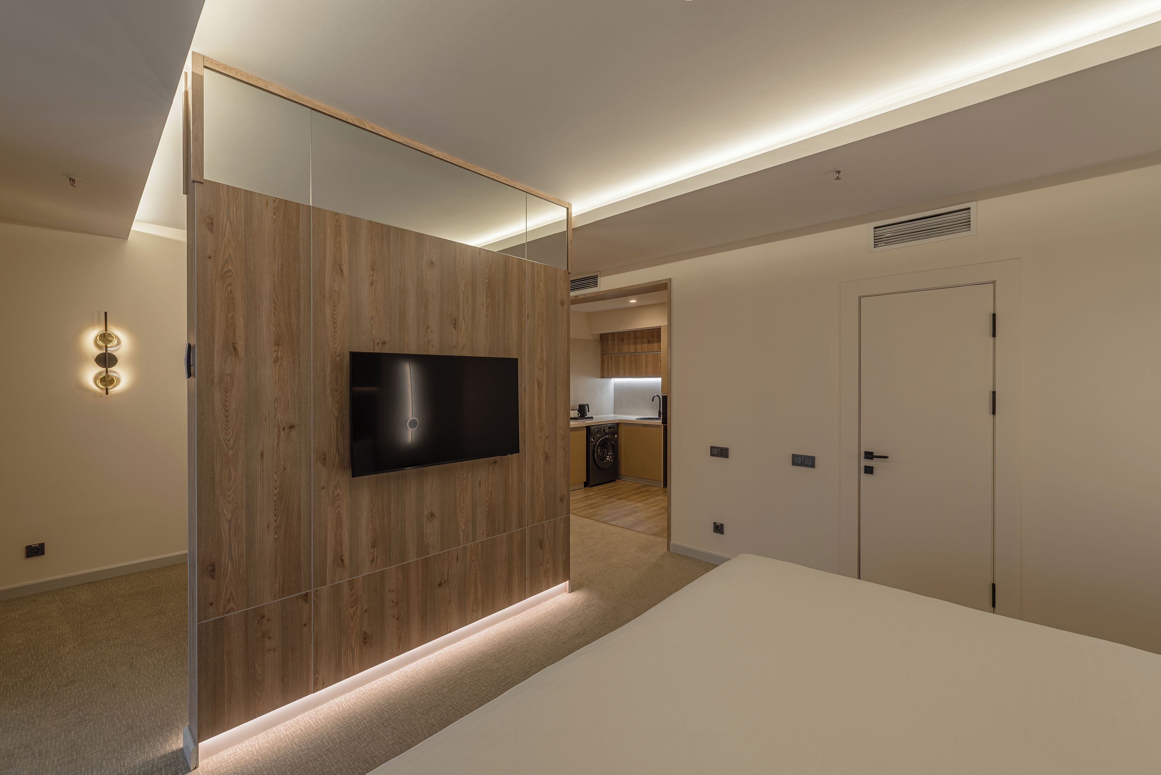 Una habitación de hotel moderna con una pared de acento de madera, un televisor de pantalla plana, una cama blanca y una zona de cocina al fondo.