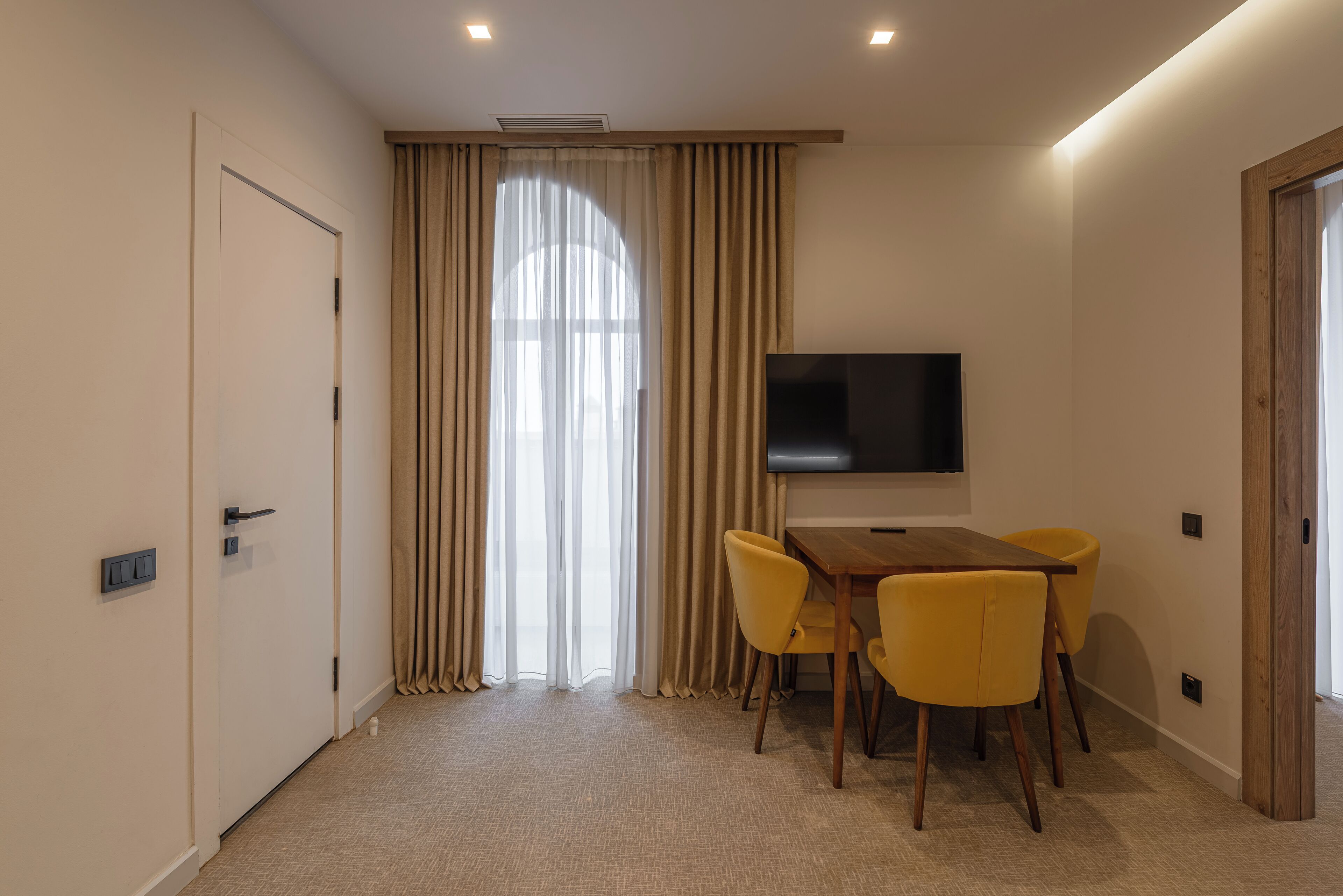 A hotel room with a large window, a flat-screen TV mounted on the wall, a wooden table with yellow chairs, and a door with a light switch.