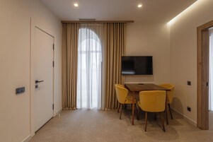 A hotel room with a large window, a flat-screen TV mounted on the wall, a wooden table with yellow chairs, and a door with a light switch.