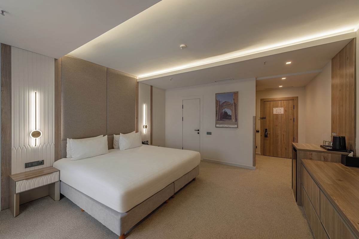 A modern hotel room with a large bed, bedside tables, a wooden wardrobe, and a desk with a coffee maker.