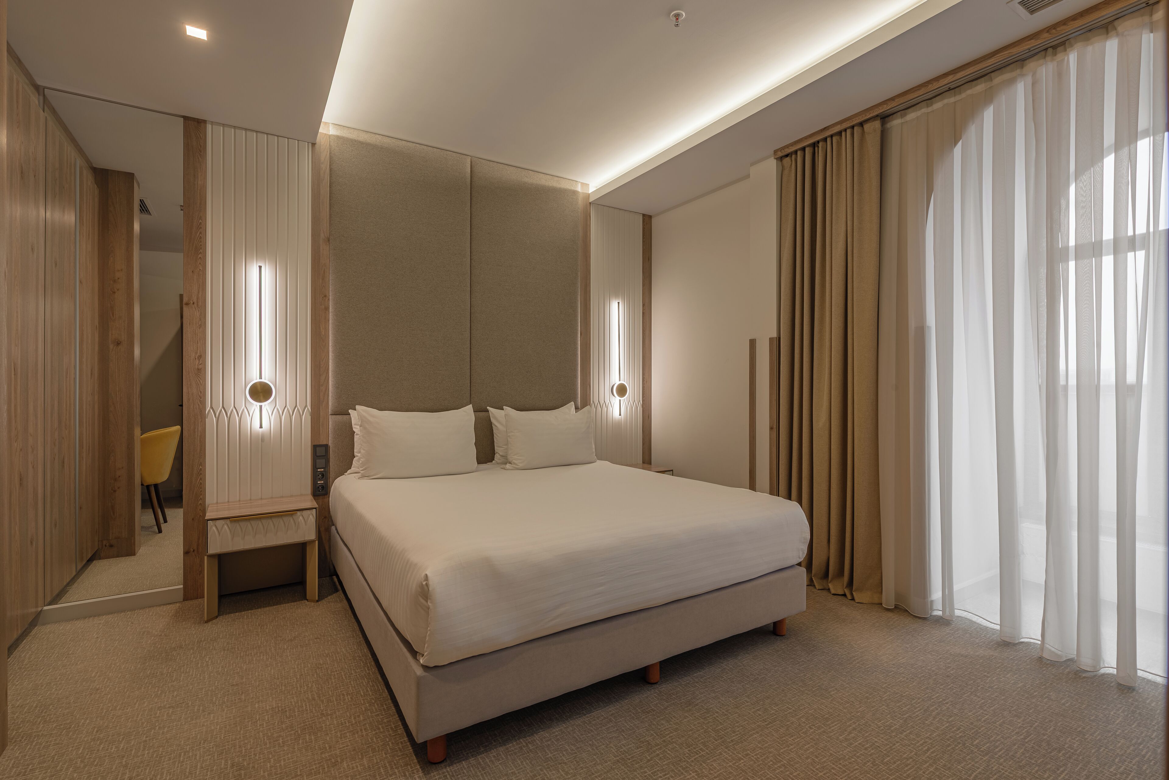 A modern hotel room with a large bed, bedside tables, a mirror, and a view of the outdoors.