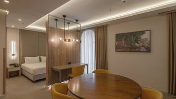Een hotelkamer met een houten bureau, een ronde eettafel met gele stoelen, een bed met wit beddengoed en een schilderij aan de muur.