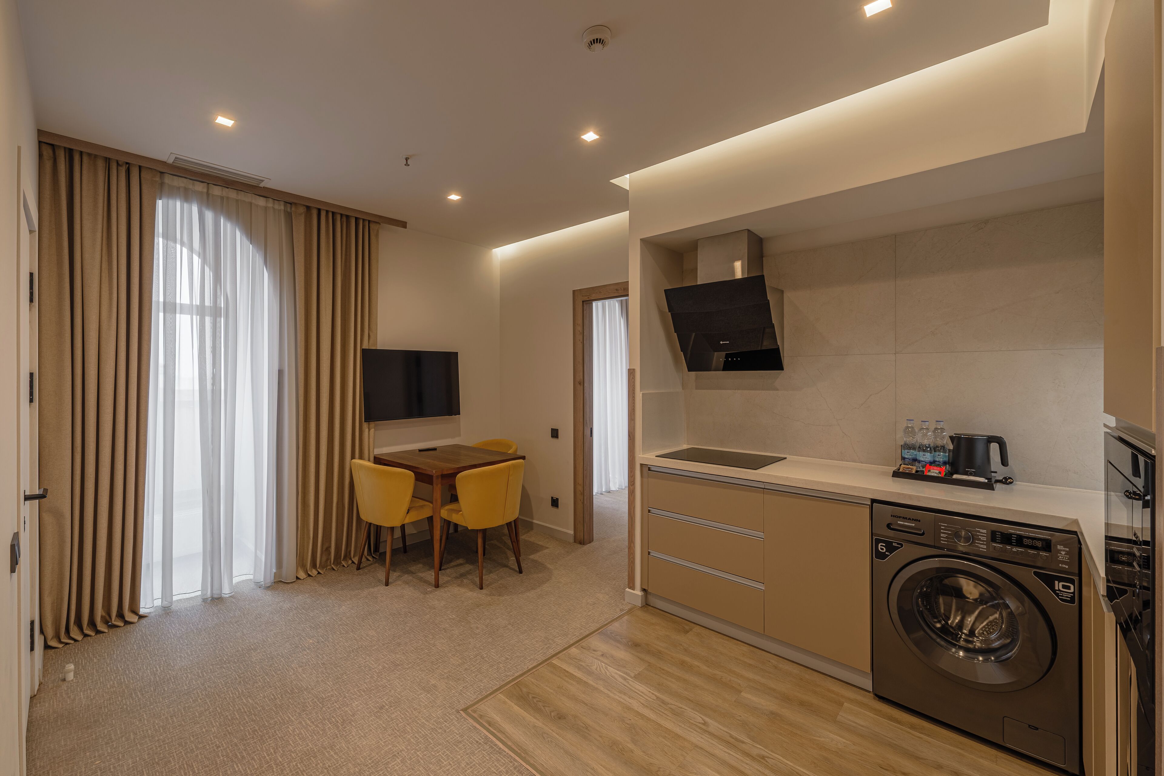 A modern kitchen with a built-in washer and dryer, a dining area with yellow chairs, and a flat-screen TV mounted on the wall.