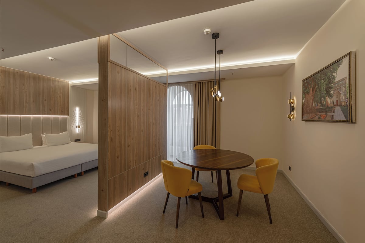 A modern hotel room with a wooden accent wall, a bed, a dining table with four chairs, and a painting on the wall.