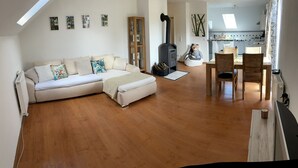 Un salon chaleureux avec un canapé d’angle, un coin repas avec des chaises en bois, une cheminée et une cuisine en arrière-plan.