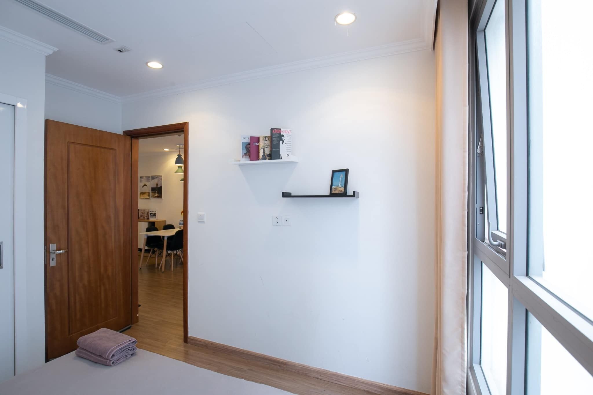 Un couloir avec une porte en bois, un mur blanc orné d’une étagère où sont posés des livres et d’un tableau encadré, et une fenêtre sur la droite.