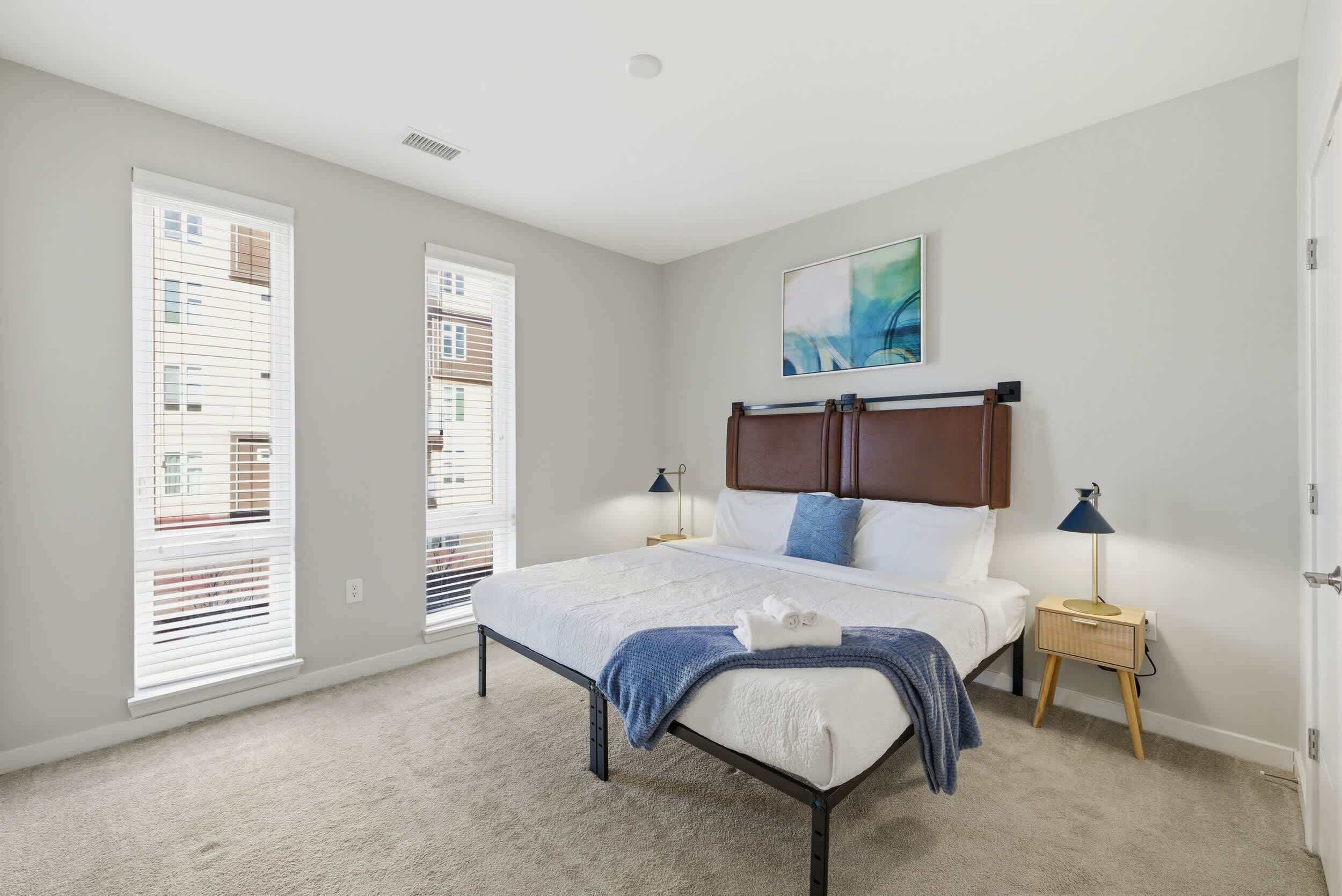 A bedroom with a bed, two bedside tables, a nightstand, a wall-mounted artwork, and large windows with blinds.