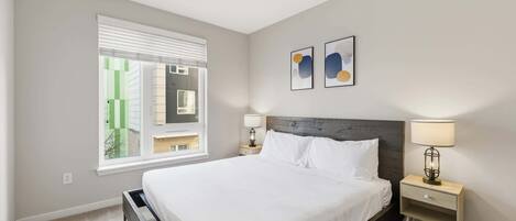 A bedroom with a large bed, a nightstand with a lamp, a window with blinds, and two wall art pieces.