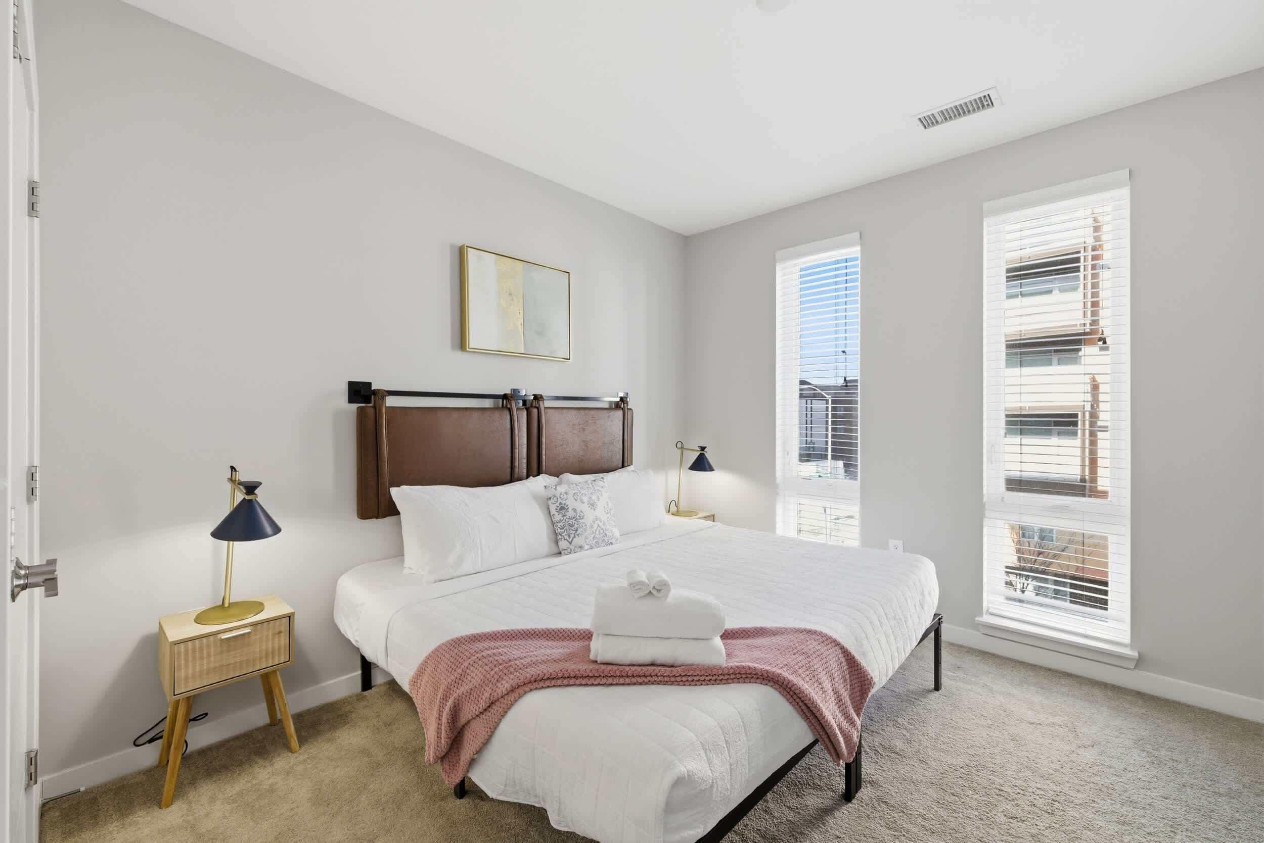 A bedroom with a bed, bedside lamps, a nightstand, a window with blinds, and a door.