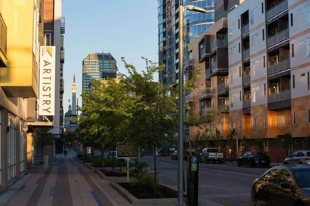 A modern urban street with multi-story buildings, a clear sky, and parked cars.