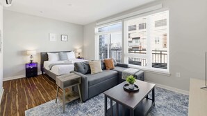 A modern hotel room with a bed, a gray sofa, a small table, and a large window with blinds. - CozySuites at Artistry Indy (Indianapolis)