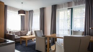 Un salon moderne avec un canapé, une table à manger et des chaises. Il y a une grande fenêtre avec des rideaux, un luminaire suspendu et une vue sur la verdure à l’extérieur.