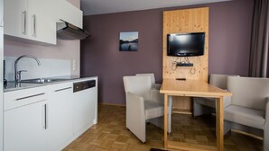 A compact kitchen with a dishwasher, a sink, and a built-in stove. There is a wooden table with two chairs and a flat-screen TV mounted on a wooden panel.