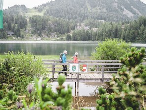 Un puente de madera sobre un lago con dos personas caminando, rodeado de vegetación y casas al fondo.