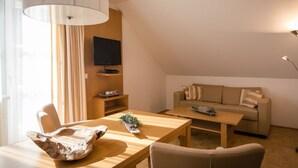 Un salon moderne avec une table à manger en bois, un canapé, un téléviseur fixé sur une étagère en bois et une grande fenêtre ornée de rideaux légers.