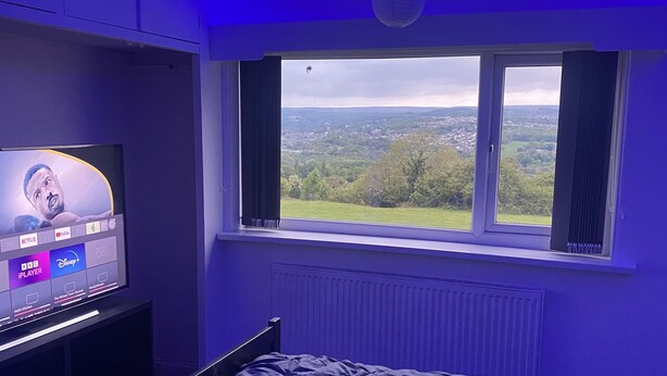Un dormitorio con cama, televisor mostrando servicios de streaming y ventana con vista a follaje.