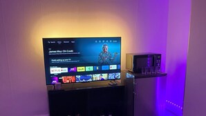 Una sala de estar con una televisión de pantalla plana montada sobre un soporte negro, un refrigerador pequeño y un ventilador de techo.