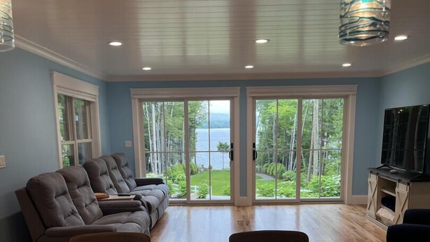 A spacious living room with a large window offering a view of a lake and forest. There are two leather sofas and a dining table with chairs. - Timeless Memories on Newfound Lake (Hebron)
