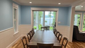 A dining room with a long wooden table and chairs, large windows with a view of greenery and water, and a ceiling with recessed lighting. - Timeless Memories on Newfound Lake (Hebron)