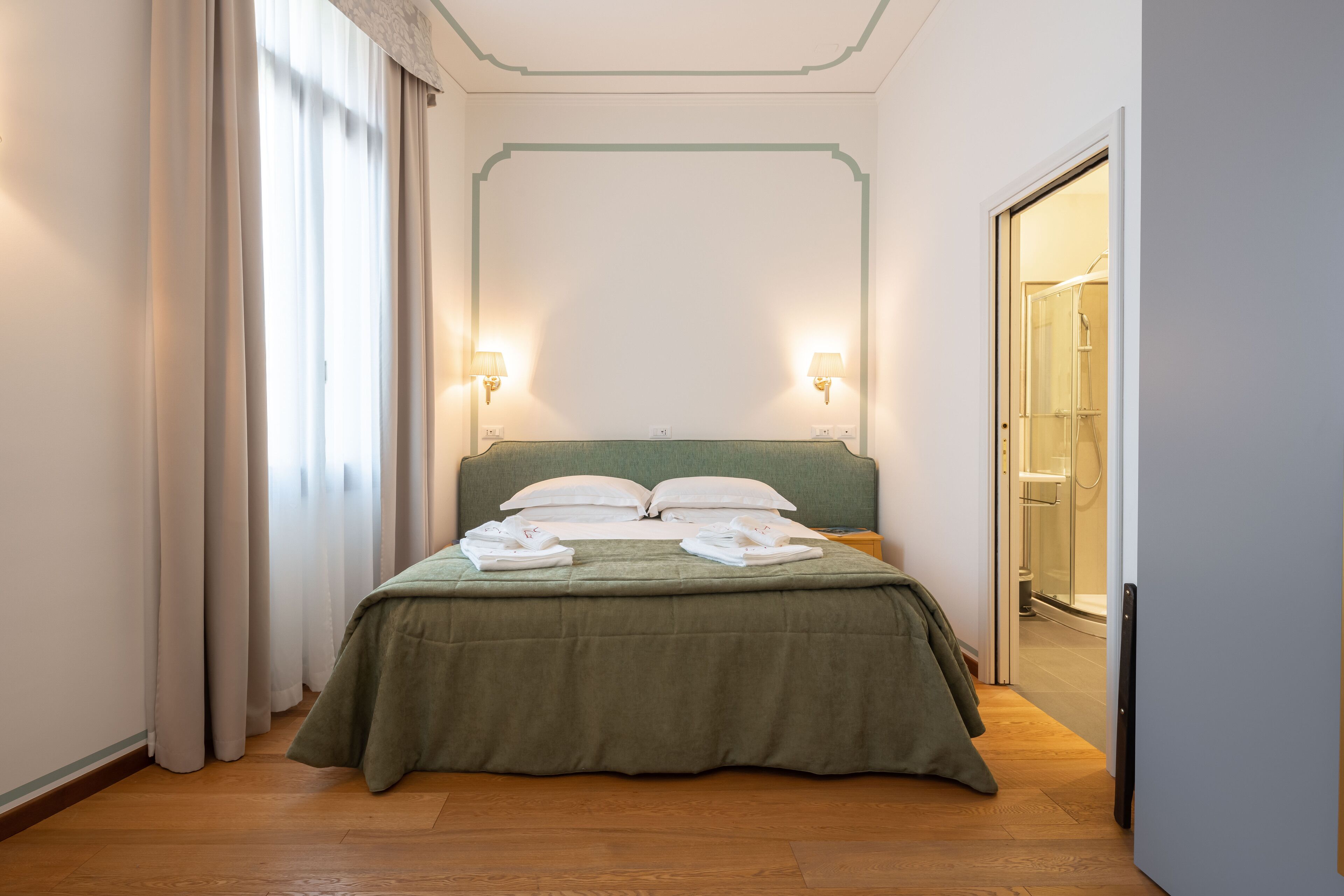 Un lit bien fait, avec une couvre-lit verte et des oreillers blancs, deux lampes fixées au mur et un plancher en bois.