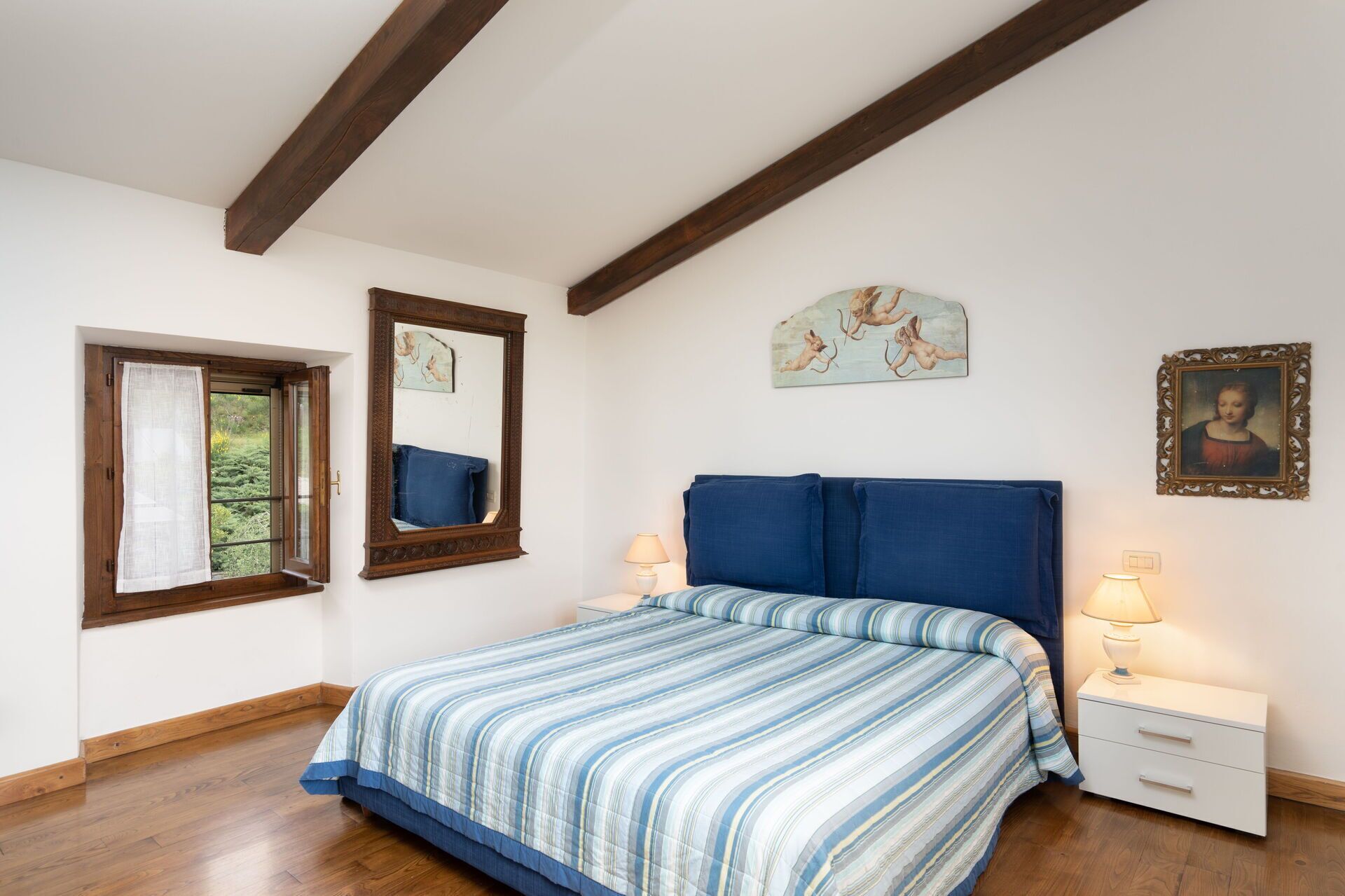 A bedroom with a bed, a nightstand, a lamp, a window with curtains, a mirror, and wall art.