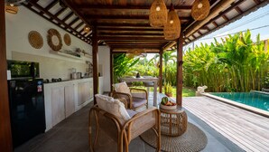 Un espacio al aire libre techado con mobiliario de mimbre, piscina y zona de cocina con armarios y refrigerador.