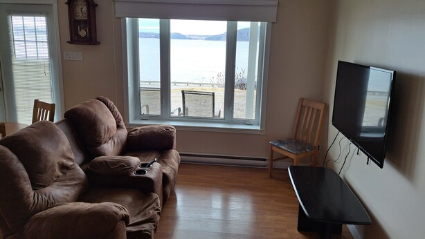 A living room with a brown leather couch, a wooden chair, a flat-screen TV mounted on the wall, and a large window with a view of a body of water and distant hills. - Very nice little condo for rent (Saguenay)