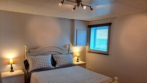 A neatly made bed with a patterned bedspread, two bedside lamps, a nightstand, and a window with blinds. - Very nice little condo for rent (Saguenay)