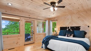 Un dormitorio de cabaña de madera con una cama, un ventilador de techo y grandes ventanas con vistas a la vegetación.