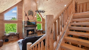 Interior de una cabaña de troncos con chimenea, escalera de madera y ventanas amplias que dan a vistas del exterior.