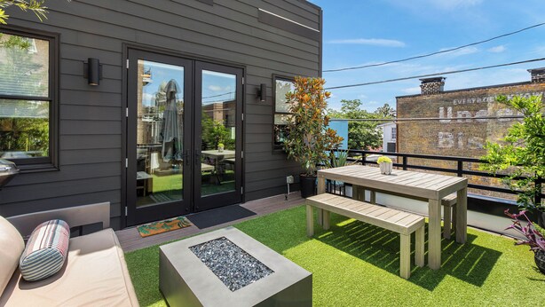 A modern outdoor patio with a wooden table and bench, a fire pit, and a view of a brick building in the background. - Modern Luxury Living in Unbeatable Location w/ Rooftop Deck by Lucky Savannah (Savannah)