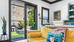 A living room with a yellow sofa, a coffee table, and a view of a brick building through a glass door. - Modern Luxury Living in Unbeatable Location w/ Rooftop Deck by Lucky Savannah (Savannah)