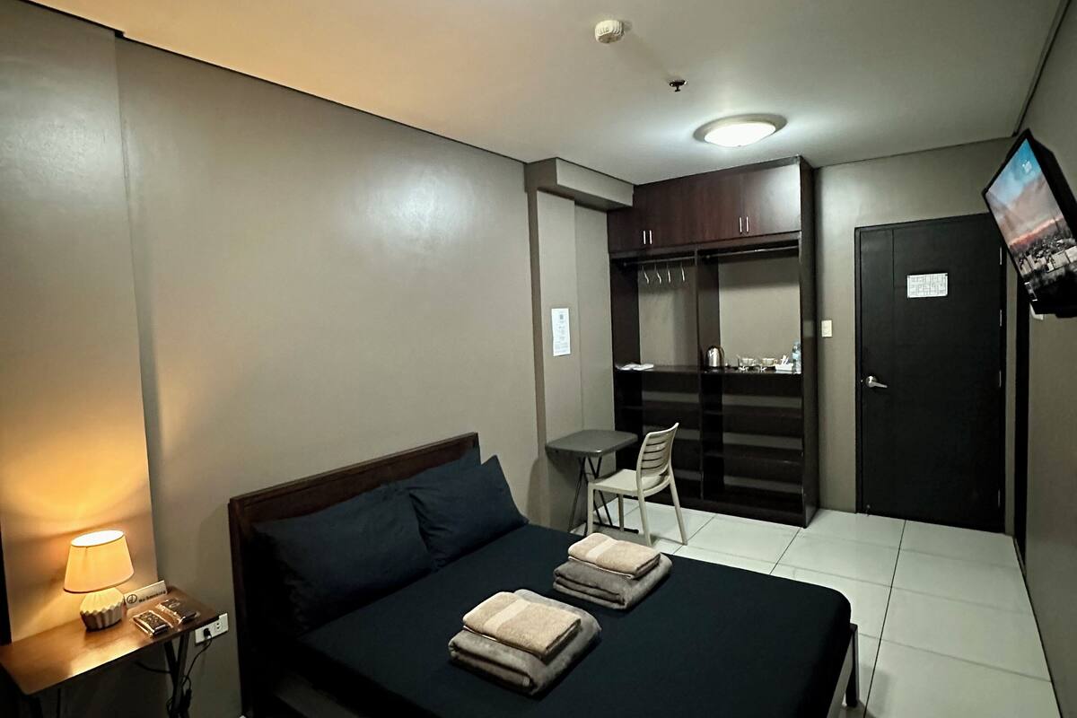 A hotel room with a bed, bedside lamp, wardrobe, and a small table with a chair.