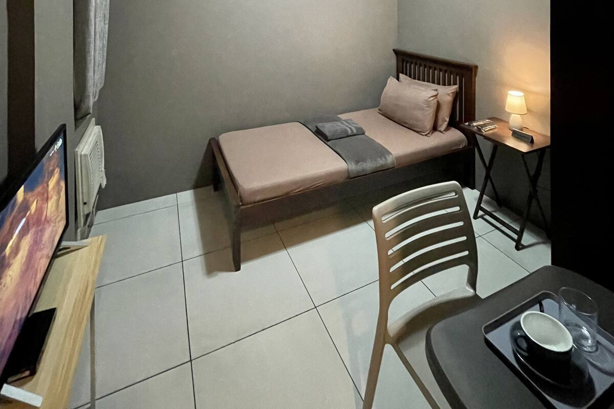 A small, compact room with a single bed, a chair, a small table with a lamp, and a TV on a stand.