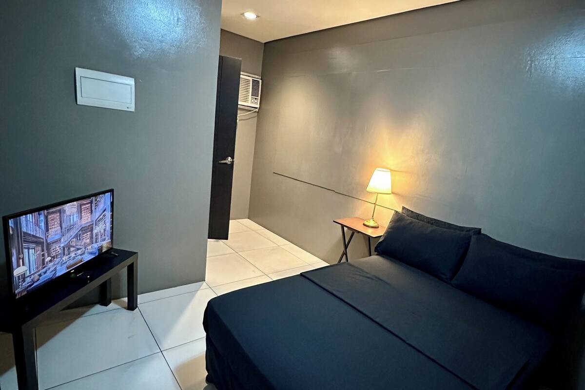 A modern hotel room with a large bed, a flat-screen TV on a stand, a bedside table with a lamp, and a wall-mounted air conditioning unit.