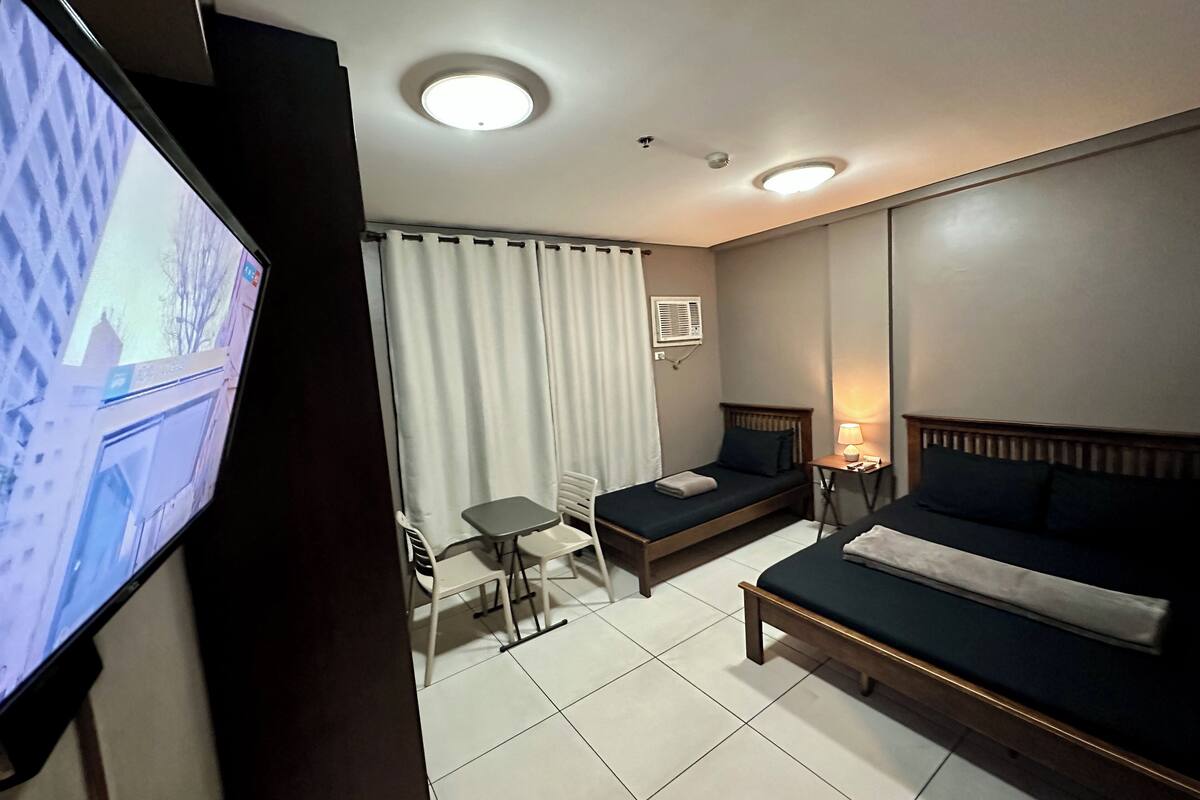 A hotel room with two single beds, a small table, a chair, a TV, and a ceiling light.