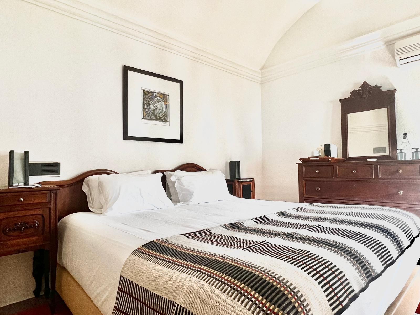 Una cama bien tendida con una colcha a rayas, un aparador de madera con un espejo y un cuadro en la pared.