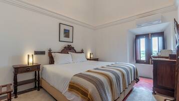 Un dormitorio con una cama grande, mesitas de noche, una mesita auxiliar, un cuadro enmarcado en la pared y una ventana con cortinas.