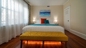 A bedroom with a bed, bedside lamps, a painting, a door, and a window with curtains. - Splendor on the Lake, 1928 Cottage-Walk Everywhere (Saint Augustine)